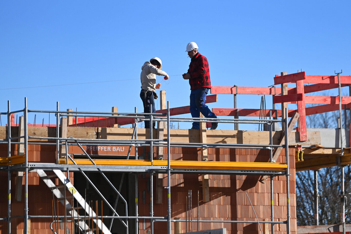 Auf einer Baustelle stehen zwei Bauarbeiter und bauen Wohnraum – finanziert auf Pump, nämlich durch die Aufhebung der Schuldenbremse – das Geld soll in alle Arten von Infrastruktur gesteckt werden